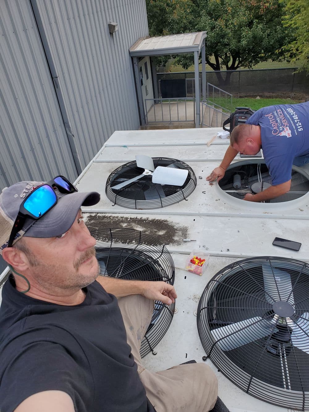 Technicians repairing rooftop air conditioning unit with fans and tools visible.