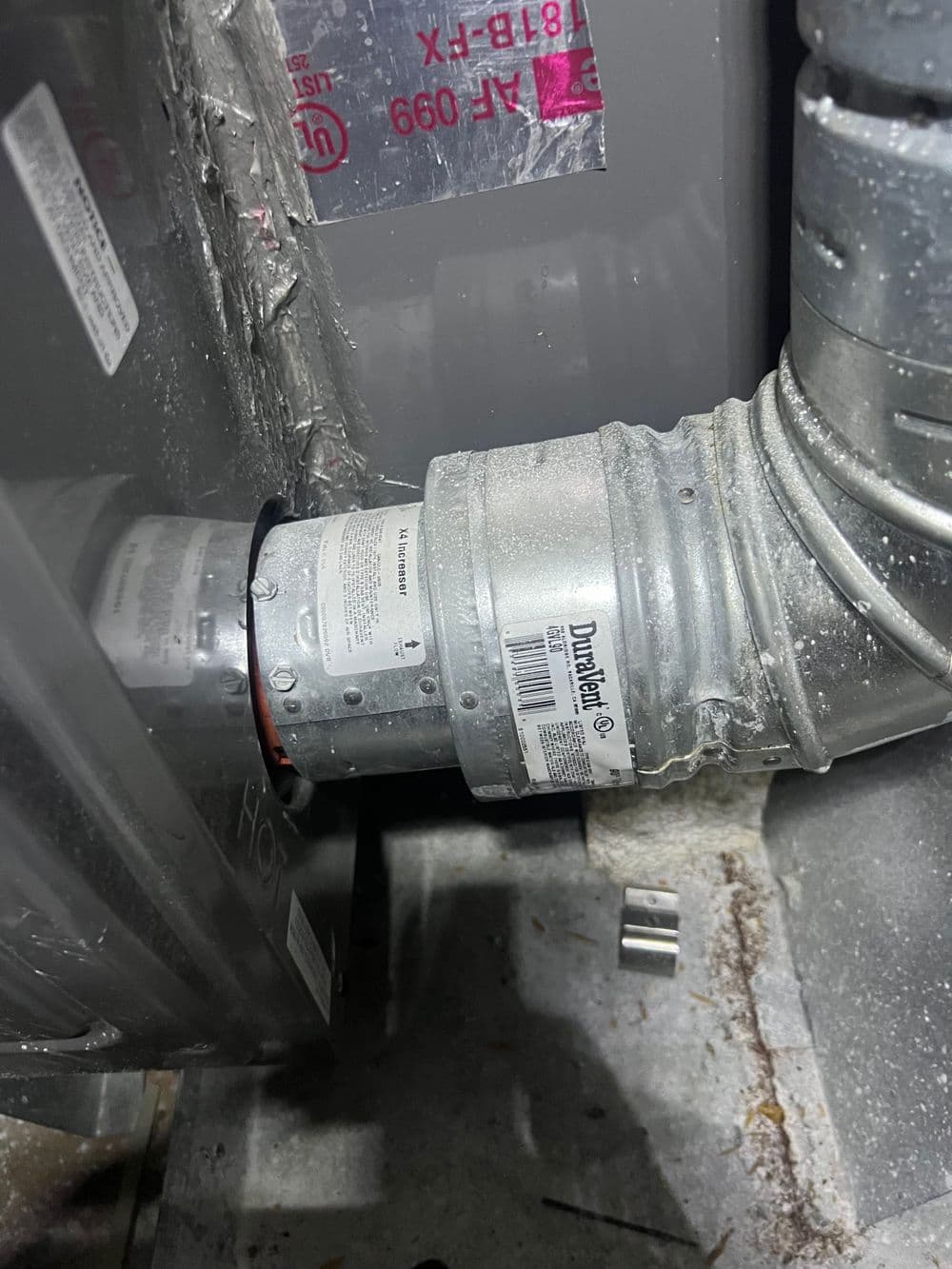 Close-up of HVAC ductwork connector with visible labels and metallic surfaces in indoor setting.