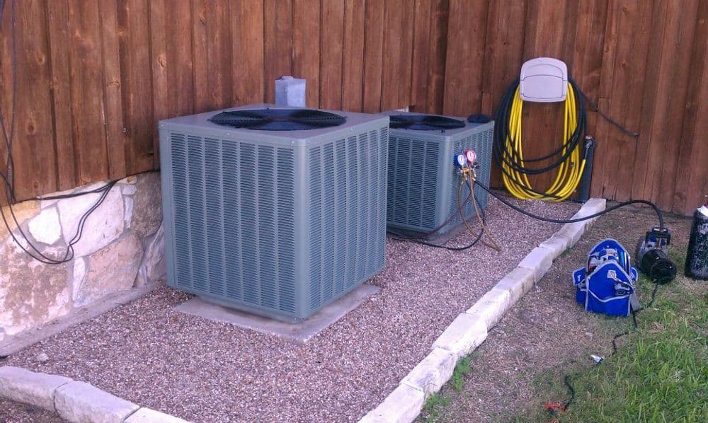 Two outdoor air conditioning units beside a wooden fence with yellow extension cords.